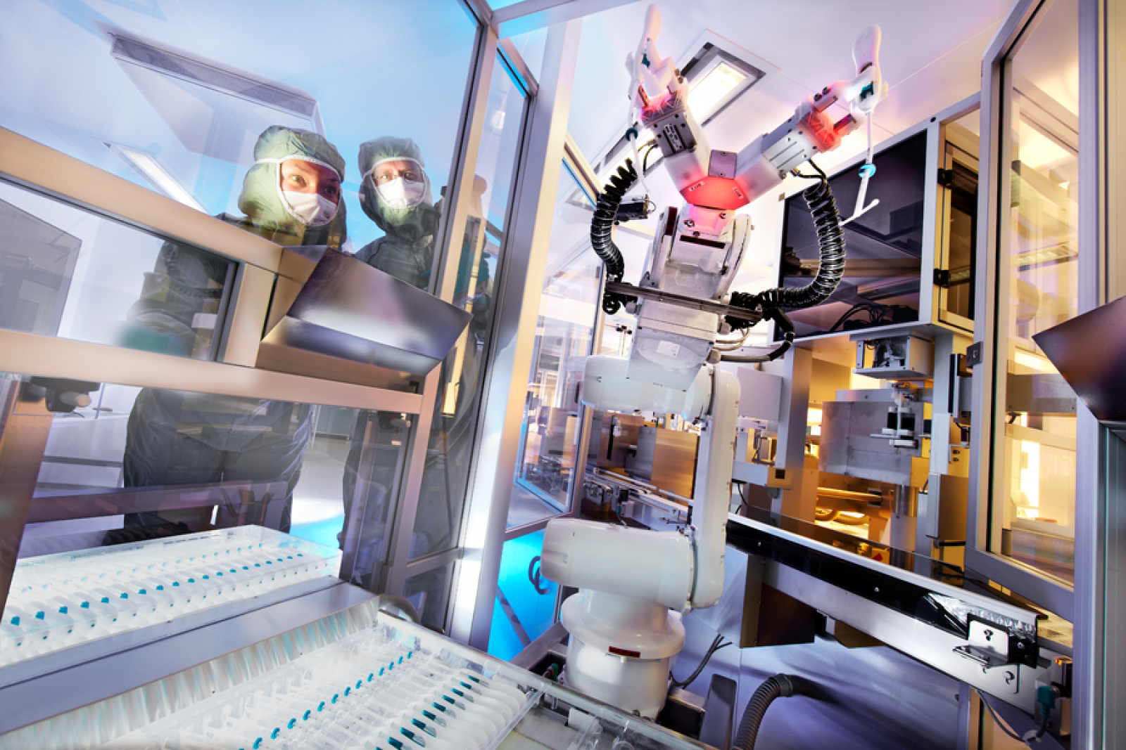 Two researchers in protective suits look through the window of a machine in a factory